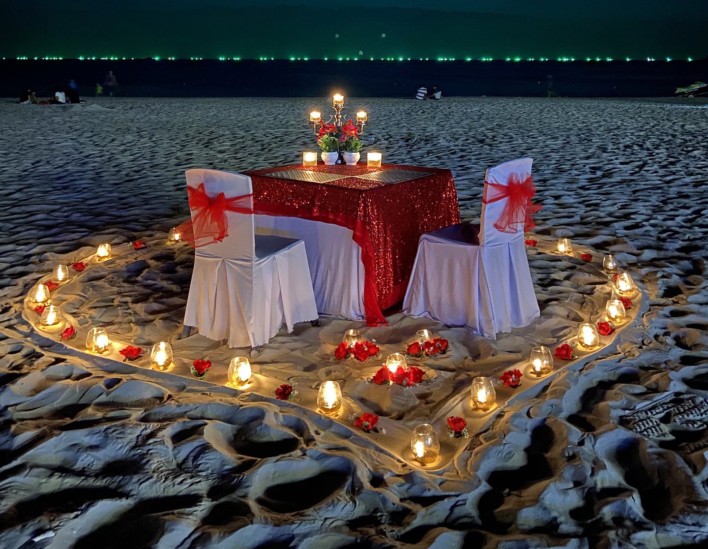 romantic candle lit dinner for 2 on a sandy beach with the table and chairs surrounded by a heart made from roses and lighted candles